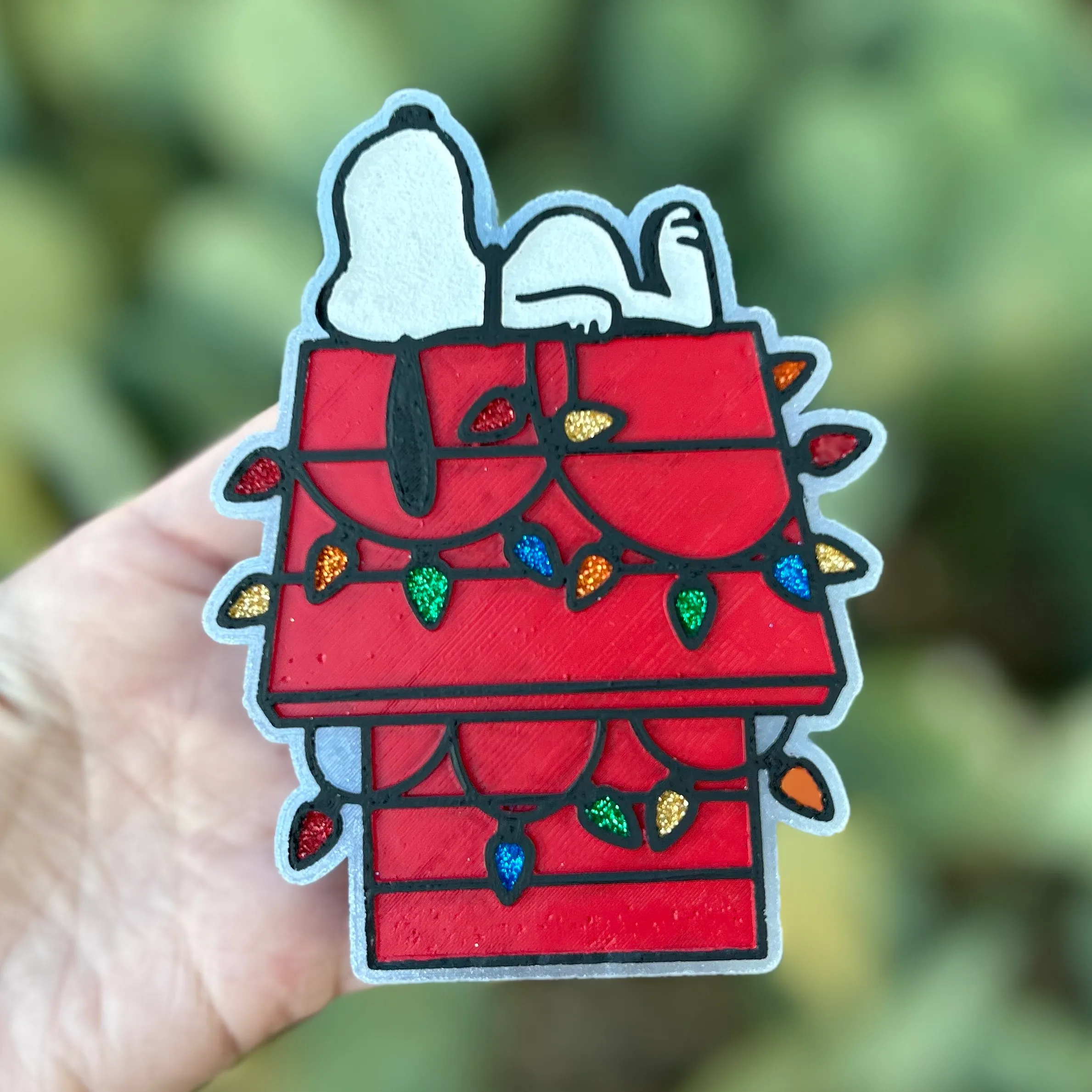 Christmas car freshie shaped like a sleeping cartoon dog on a red doghouse draped with colorful glitter Christmas lights in a stained-glass style, held in hand against greenery.