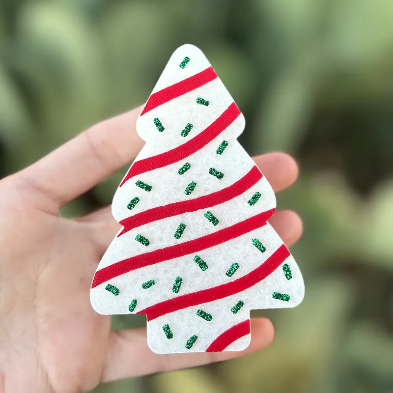 Christmas tree shaped car freshie made to look like a snack cake with white frosting, red icing stripes, and green glitter sprinkles, held in hand against a green background.