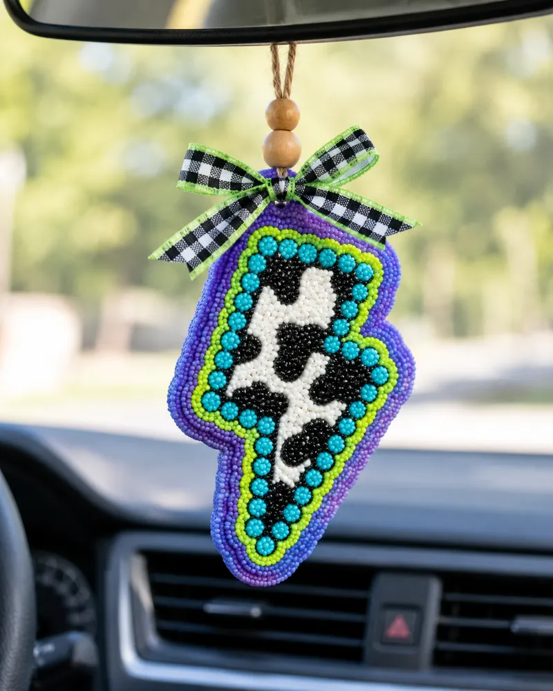 Handmade lightning bolt shaped car freshie in black and white cow print, a scented aroma bead air freshener hanging from a car rearview mirror.