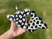 Handmade scented aroma bead car freshie shaped like a black-and-white cowhide steer head with ears up on a cream base, a western farmhouse air freshener on a cow-print beaded cord.