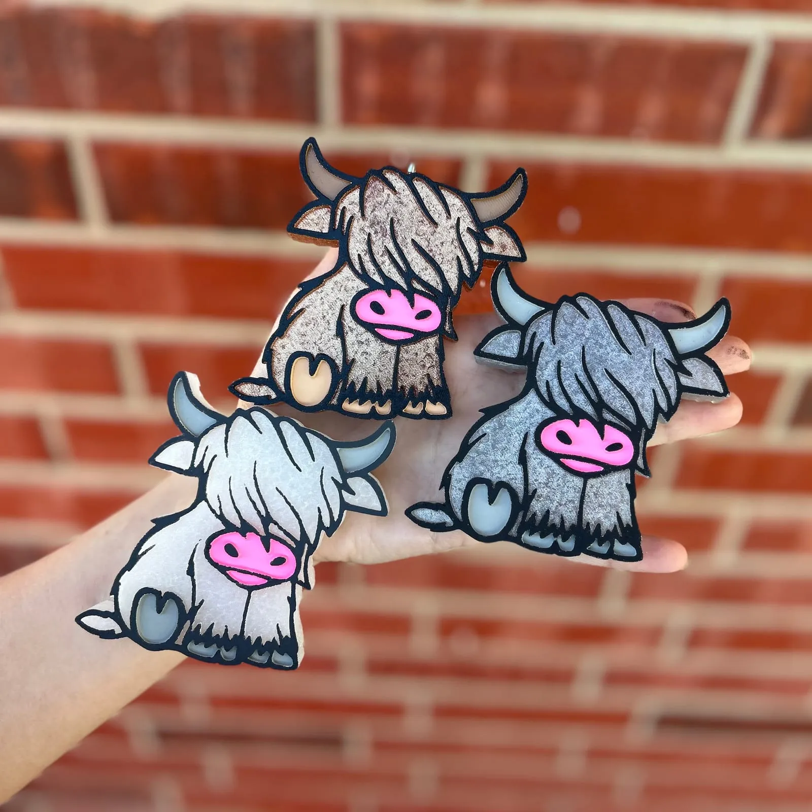 Handmade scented aroma bead car freshie air freshener shaped like a fluffy baby Highland cow calf with shaggy hair, little horns, a heart-shaped nose, and tiny hooves, shown in brown, grey, and white.
