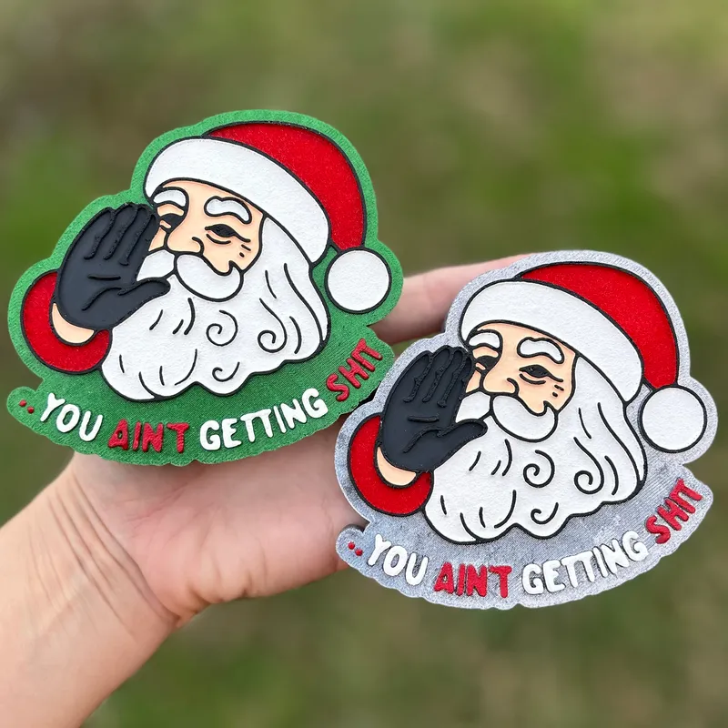 Two funny Christmas car freshies held in a hand, each showing a cartoon Santa raising his palm with the caption "You Ain't Getting Sh*t" - one on a green background and one on a silver glitter background.