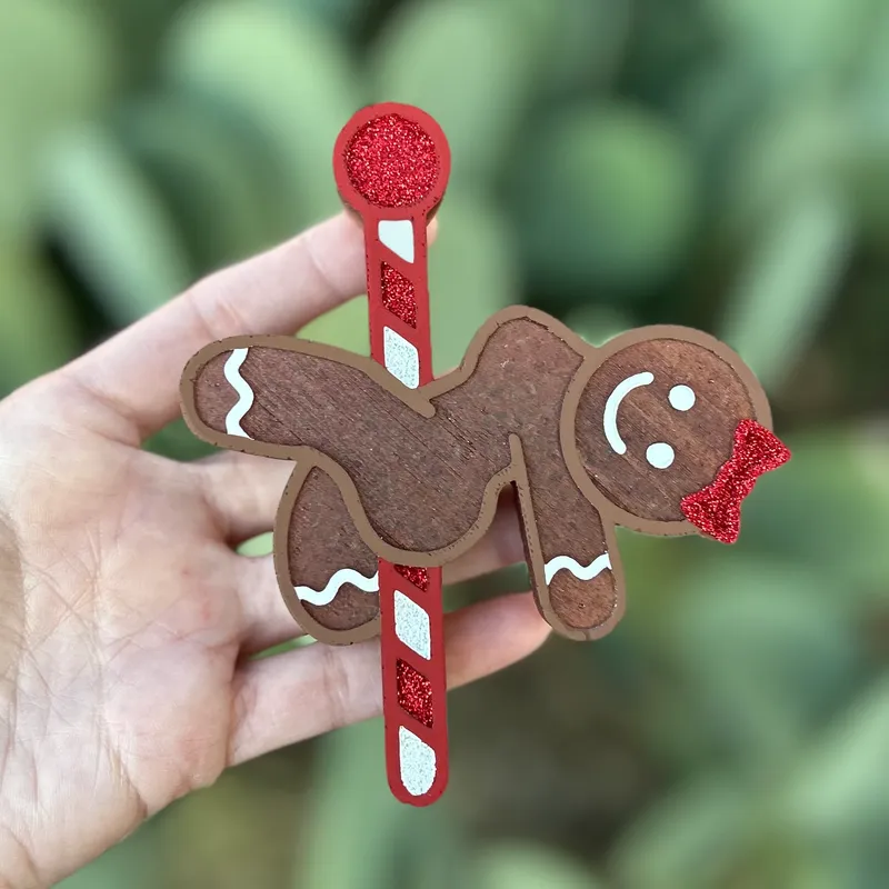 Handmade gingerbread girl Christmas car freshie with a red glitter bow, white icing details, and a candy cane stick, held in hand against a green background.