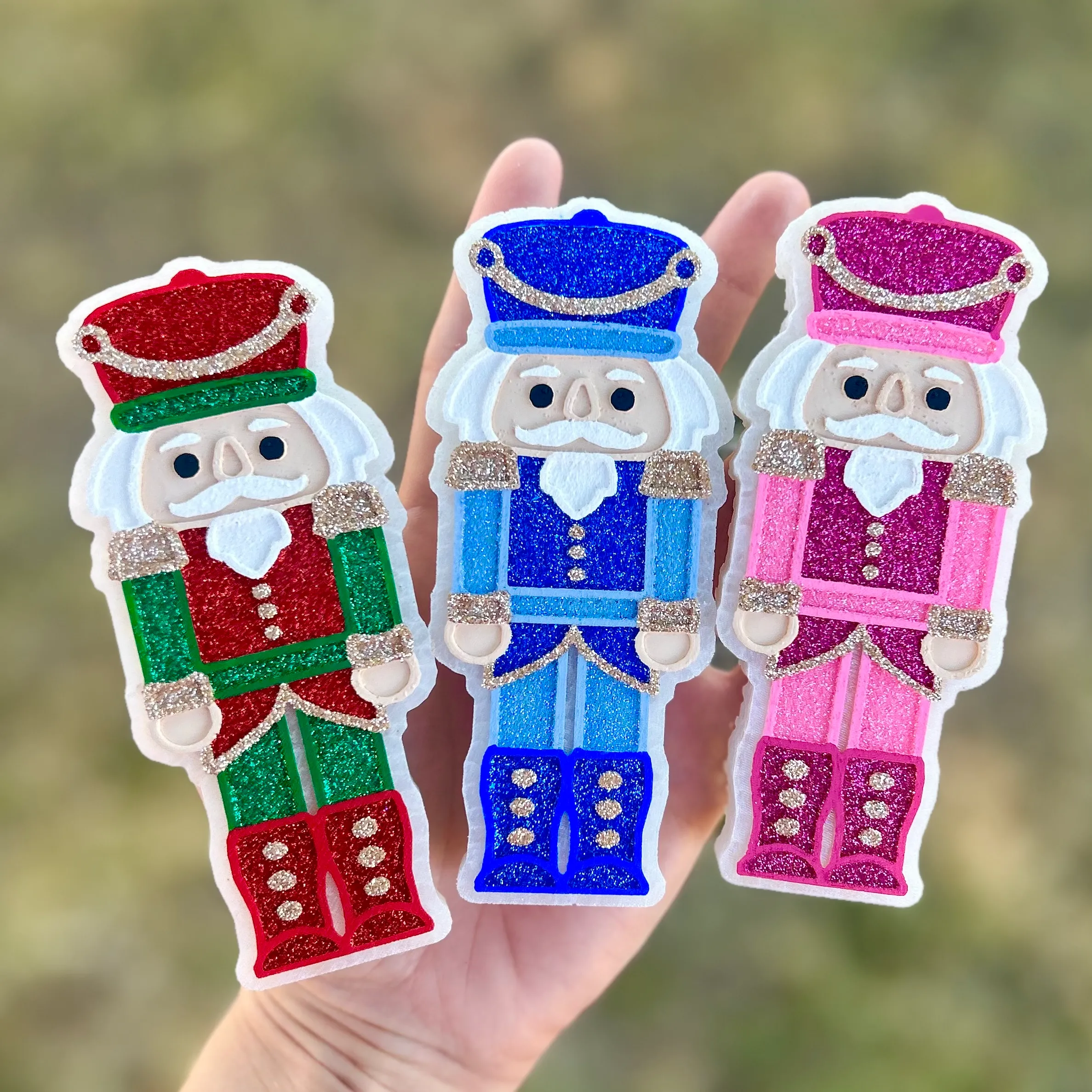 Three glitter Nutcracker Soldier car freshies held in a hand, in red and green, royal blue, and pink color variations, each a scented Christmas car air freshener.