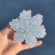 Frosted white snowflake-shaped car freshie covered in iridescent silver and blue glitter, held in a hand against a soft blue background.