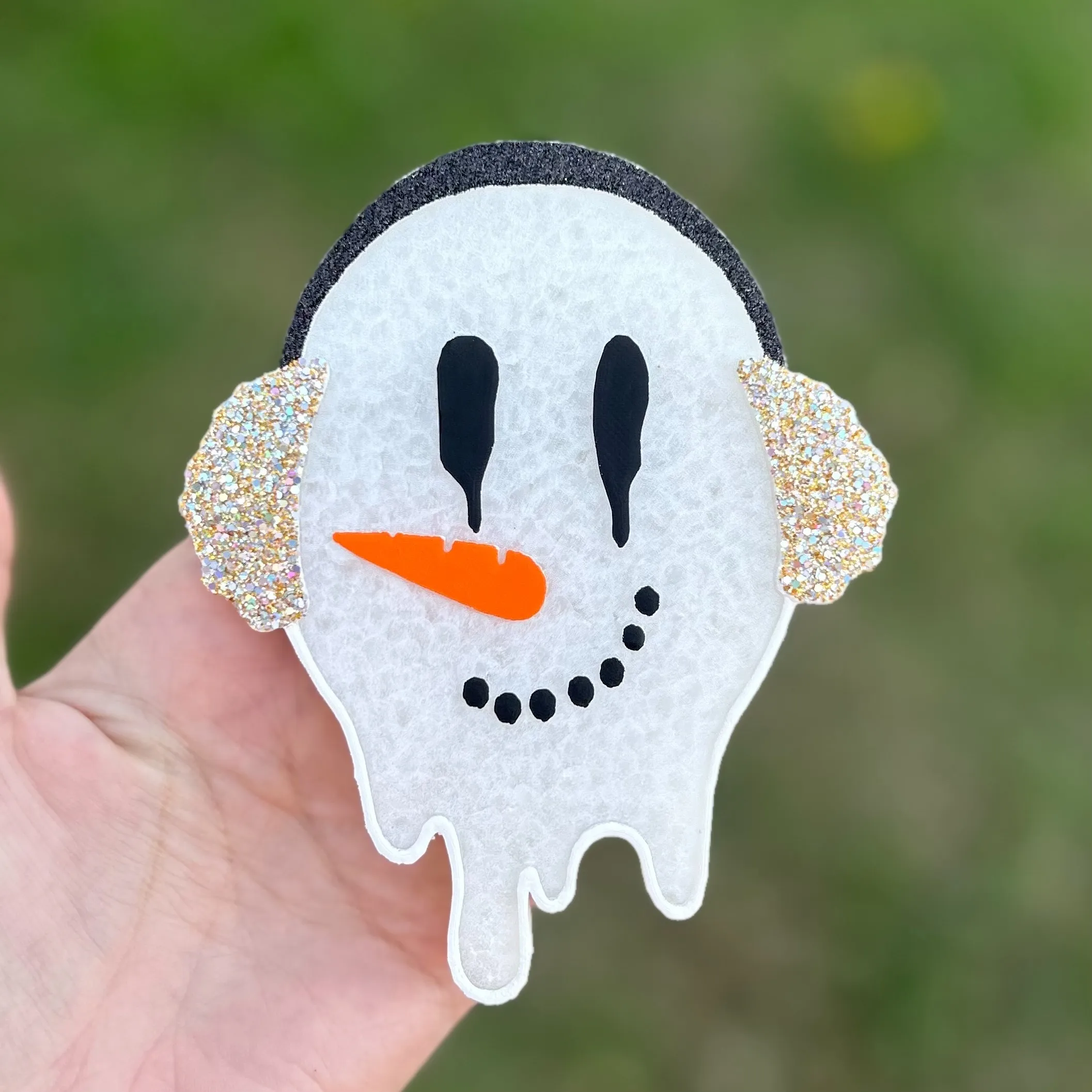 White melting snowman car freshie with sparkly iridescent glitter earmuffs, black drippy eyes, orange carrot nose, and a black dot smile, held against a green outdoor background.